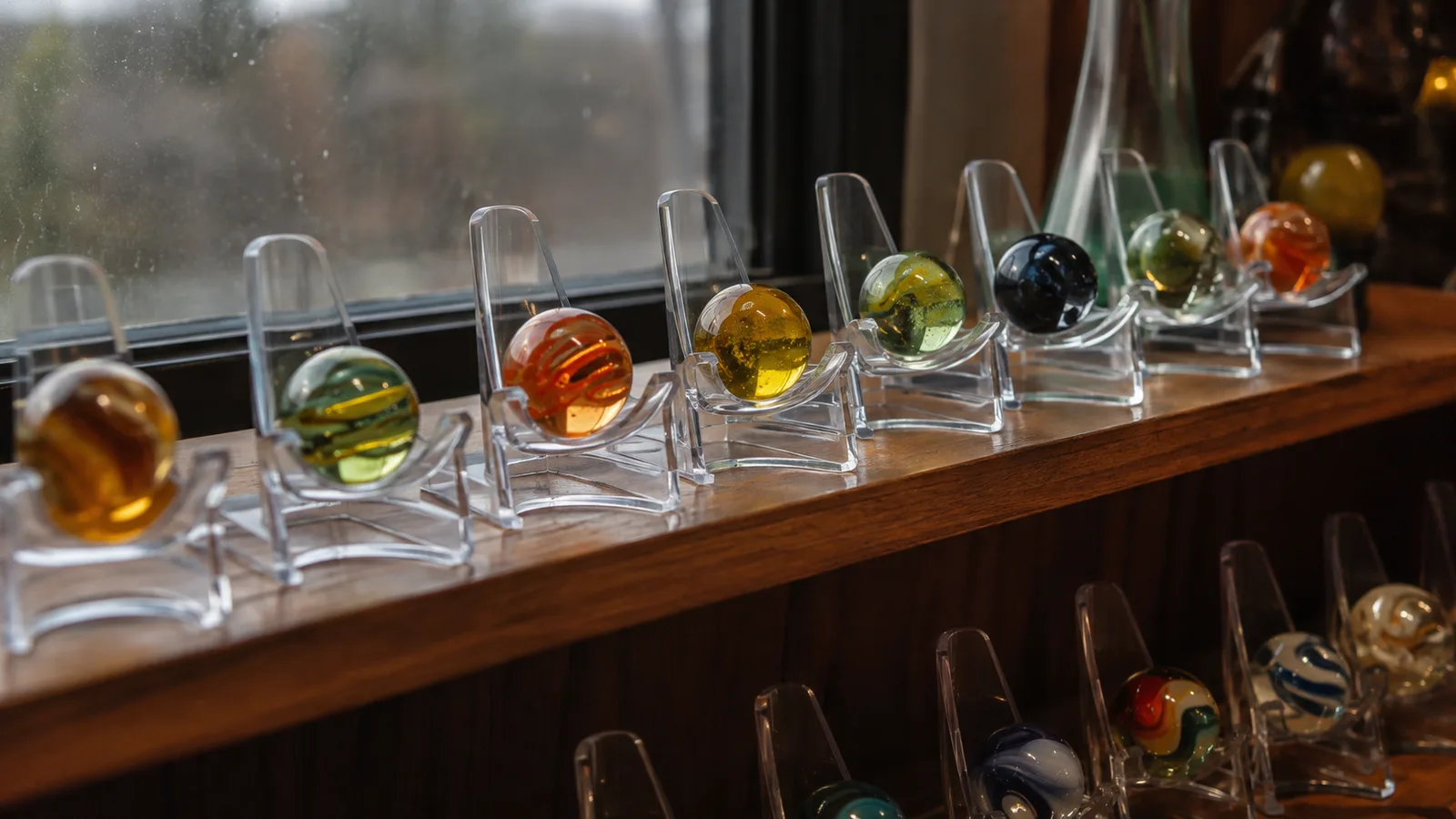 A row of clear acrylic easel stands on a shelf, each holding a different marble, with warm afternoon light passing through the glass