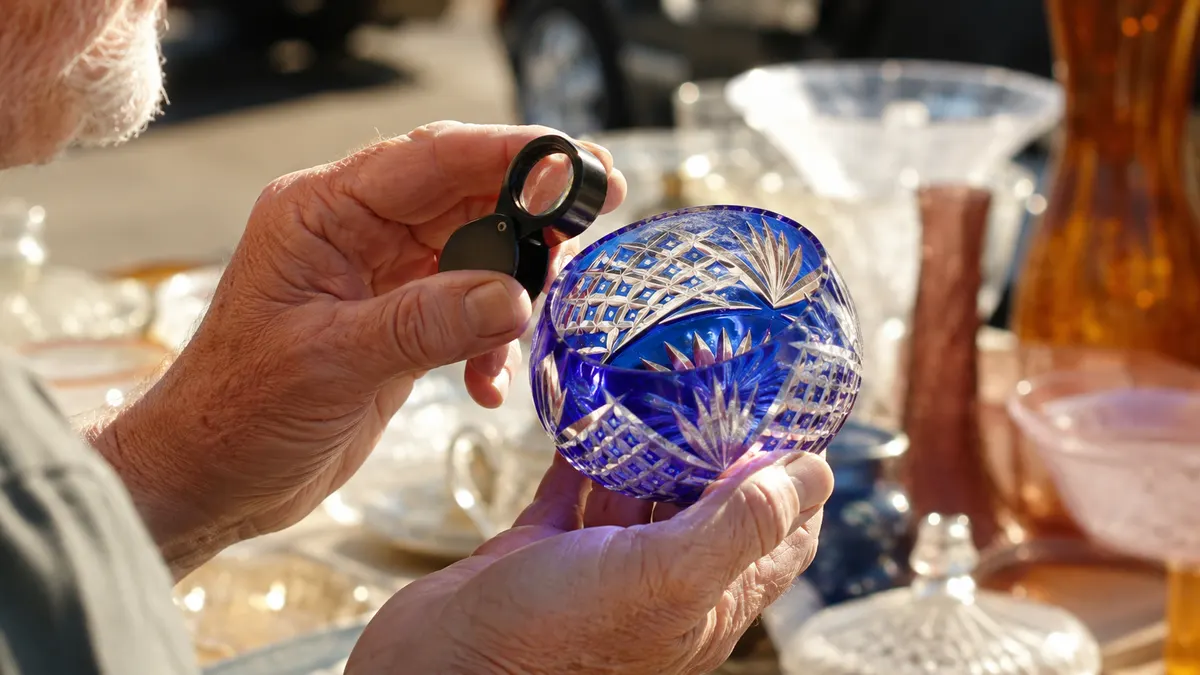 An older man's hands holding open a small folding jeweler's loupe close to a cut-glass sugar bowl on a dealer's card table at an outdoor market