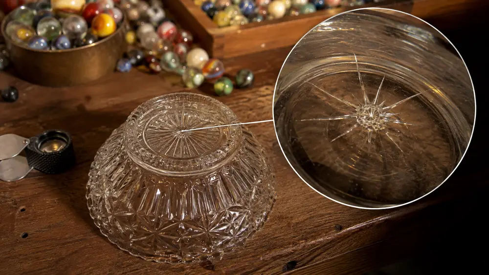 A close view of an antique pressed-glass bowl's raised foot with a magnified inset showing a small star crack at the base mark