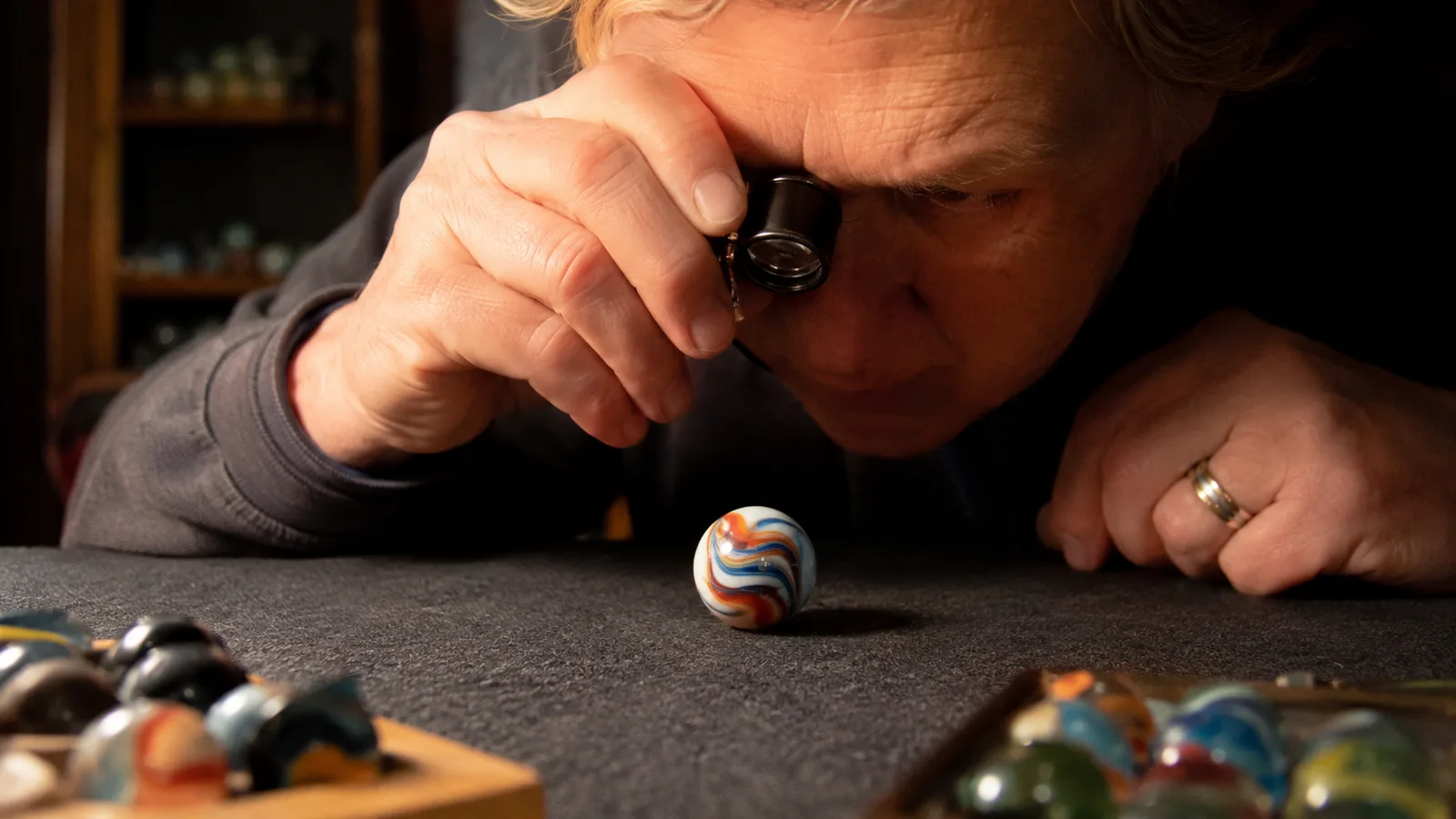 Older man's hand holding a small black jeweler's loupe up to his eye while inspecting a large glass marble on a felt pad